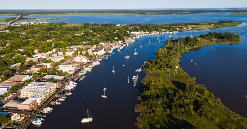 Aerial view of a river winding through a town with boats, greenery, and a blue sky in the background.