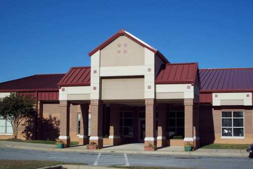 A brick building with a red metal roof and a central entrance, surrounded by green grass and blue sky.
