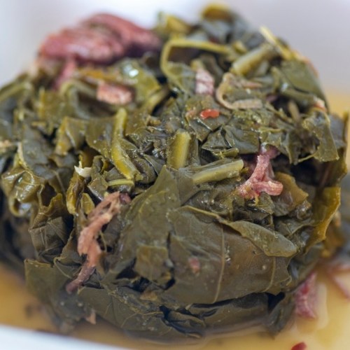 A close-up of cooked collard greens with bits of meat, served in a light broth.