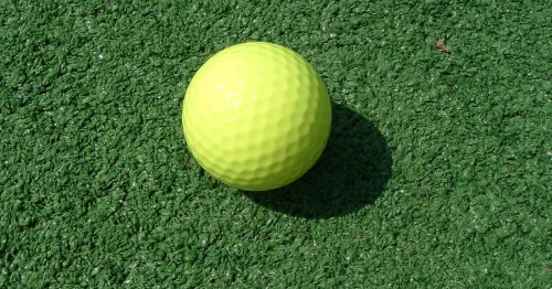 A bright yellow golf ball resting on a green artificial turf surface.