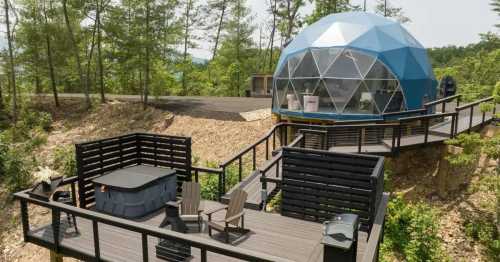 A modern geodesic dome home surrounded by trees, featuring a deck with seating and a hot tub.