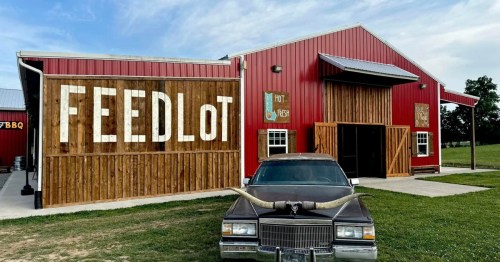 A rustic red barn with wooden accents, featuring the word "FEEDLOT" and a vintage car with longhorns in front.