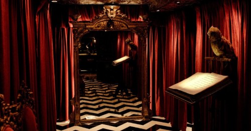 A dimly lit room with red curtains, a large ornate mirror, and a book on a stand, featuring a black and white chevron floor.