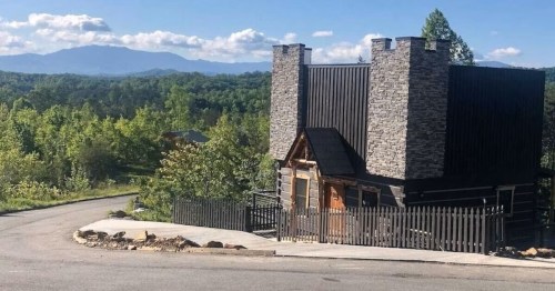 A unique house with stone accents and a black exterior, surrounded by trees and mountains in the background.