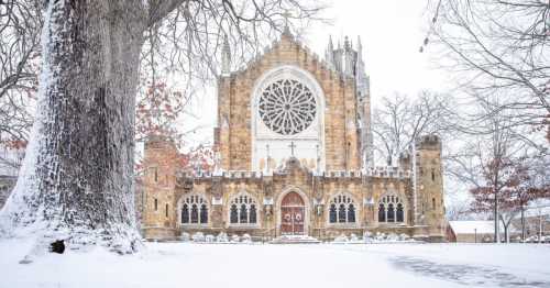 A snow-covered church with a large rose window, surrounded by bare trees and a serene winter landscape.