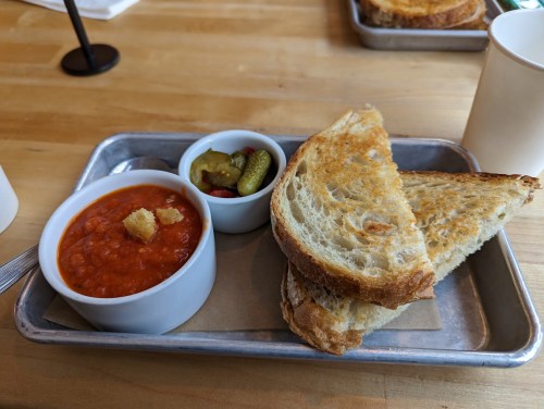 A metal tray with a bowl of tomato soup, croutons, pickles, and two slices of toasted bread.