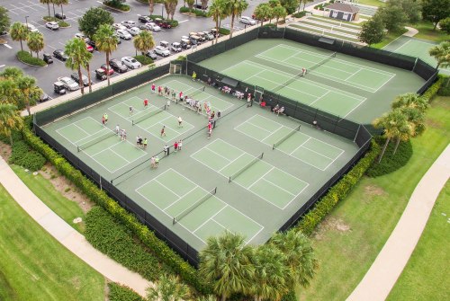 Aerial view of multiple tennis courts with players and spectators, surrounded by palm trees and parked cars.