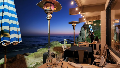 Cozy seaside restaurant at dusk, featuring outdoor heaters, tables, and a view of the ocean under a twilight sky.