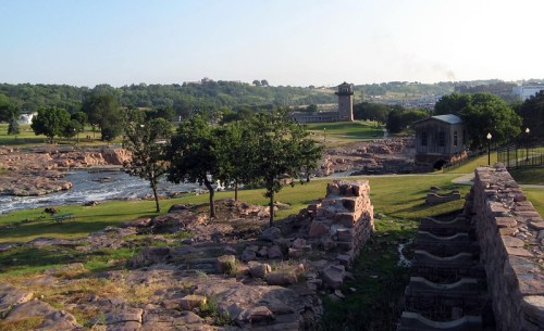 A scenic view of a park with rocky terrain, trees, and a river, featuring historic buildings in the background.