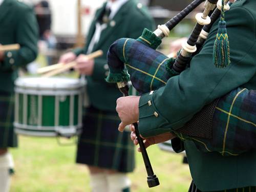 A close-up of a piper in a green uniform playing bagpipes, with drummers in the background.