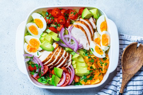 A colorful salad with sliced chicken, boiled eggs, tomatoes, cucumbers, and red onions in a white dish.