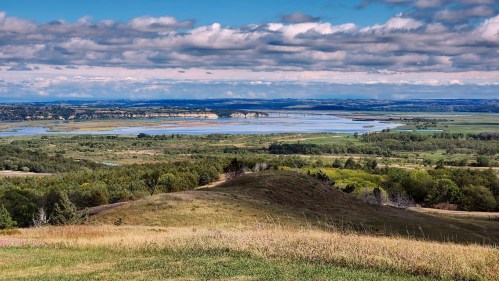 A scenic view of rolling hills and a river, with lush greenery and a cloudy sky in the background.
