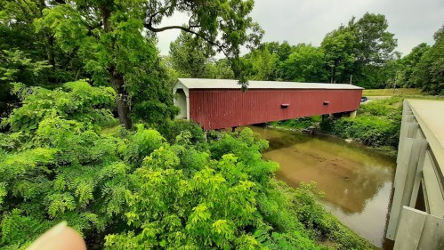 A red covered bridge surrounded by lush green trees, spanning a calm creek under a cloudy sky.