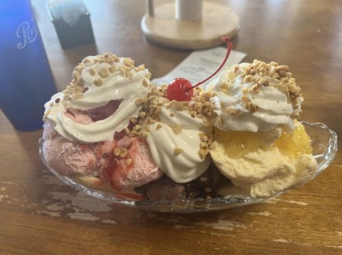 A colorful ice cream sundae with three scoops, whipped cream, chopped nuts, and a cherry on top, served in a glass dish.