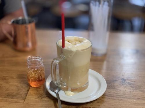 A frothy root beer float in a glass mug with a red straw, next to a small jar of syrup on a wooden table.