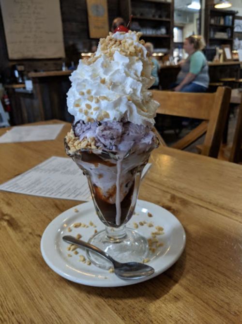 A tall ice cream sundae topped with whipped cream, nuts, a cherry, and chocolate sauce, served in a glass dish.