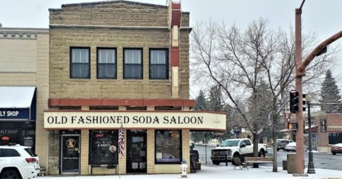 Historic building with "Old Fashioned Soda Saloon" sign, snowy street, and parked cars in a small town setting.