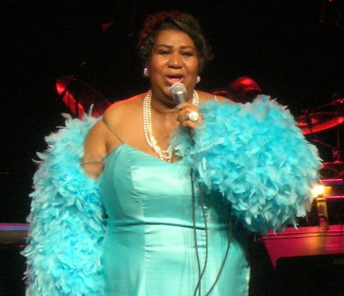 A woman in a turquoise gown and feather boa sings into a microphone on stage, surrounded by musicians.