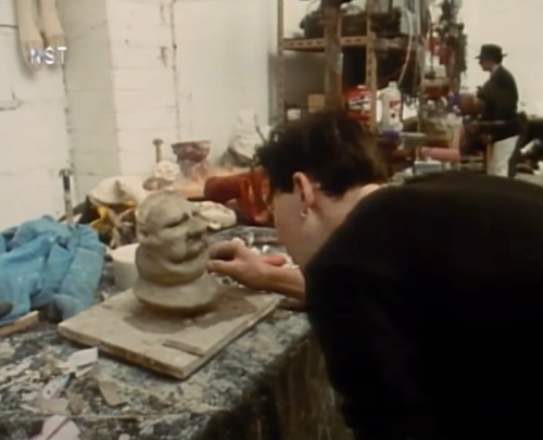 A sculptor works on a clay bust in a cluttered studio, with another person in the background.