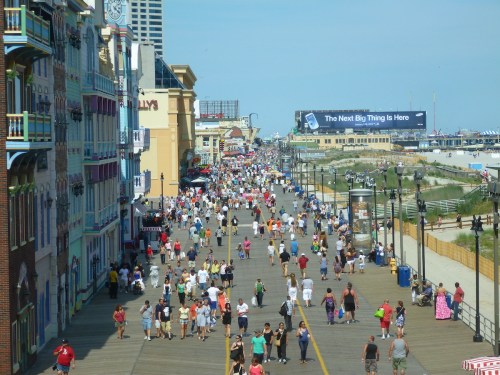 A busy boardwalk filled with people, lined with colorful buildings and a large billboard in the background.