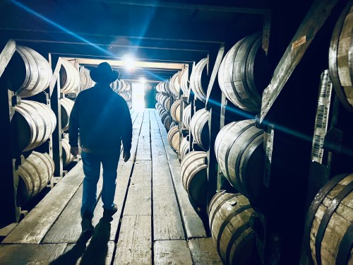 A person in a cowboy hat walks through a dimly lit storage area lined with wooden barrels.