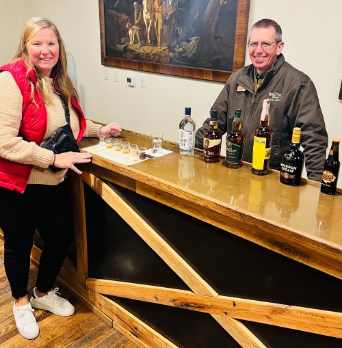 A woman and a man stand at a wooden bar, sampling spirits with bottles displayed behind them.