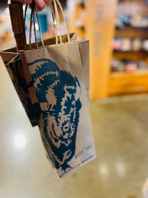 A hand holds a brown paper bag featuring a blue buffalo design, with a store interior blurred in the background.