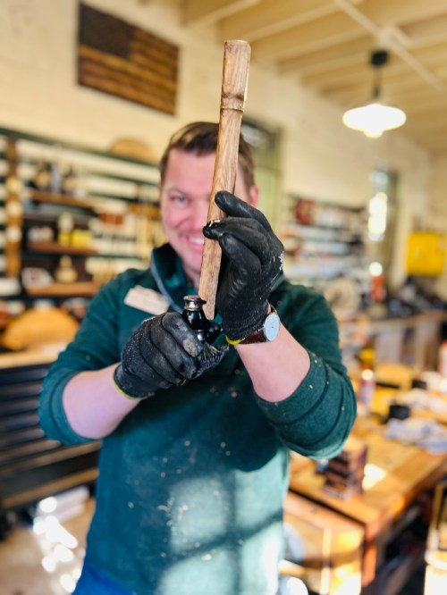 A person in a workshop holds a wooden tool, wearing gloves and smiling, with tools and shelves in the background.
