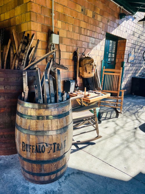 A wooden barrel labeled "Buffalo Trace" beside a table with tools and a rocking chair in a rustic outdoor setting.