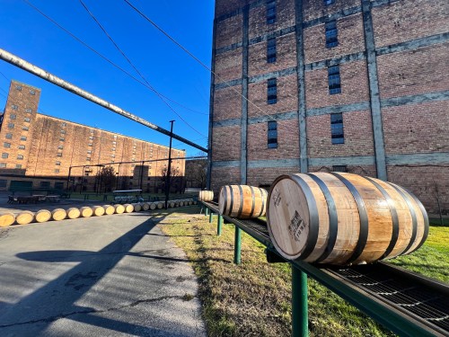 Wooden barrels lined up outside a brick building under a clear blue sky.