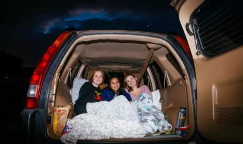 Three friends relax in the back of a car, cozy in blankets, under a night sky with clouds.