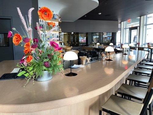 A modern bar with a curved wooden counter, stylish lamps, and a vibrant floral arrangement.