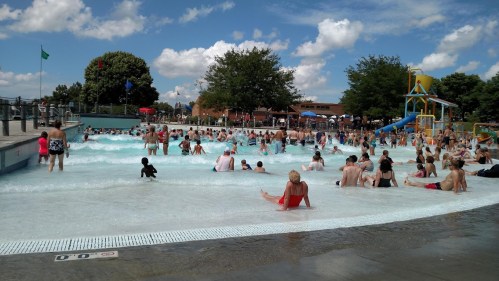 A crowded wave pool on a sunny day, with people enjoying the water and a playground in the background.