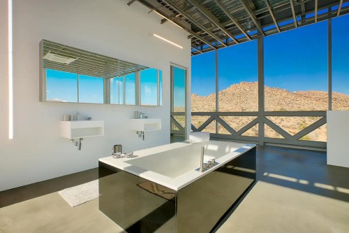 Modern bathroom with a freestanding tub, dual sinks, large mirrors, and a view of desert mountains through large windows.