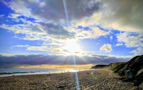 A beach scene at sunset with clouds, sun rays shining through, and people walking along the shore.