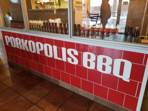 Sign for "Porkopolis BBQ" on a red tiled wall, with various sauces displayed behind a glass window.