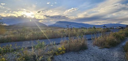 A scenic landscape featuring mountains, a path, and sun rays shining through clouds at sunset. Wildflowers in the foreground.