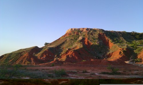 A rugged red rock formation under a clear blue sky, with green vegetation on the slopes.