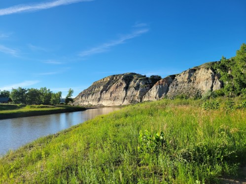 A serene riverbank scene with lush green grass and a rocky cliff under a clear blue sky.