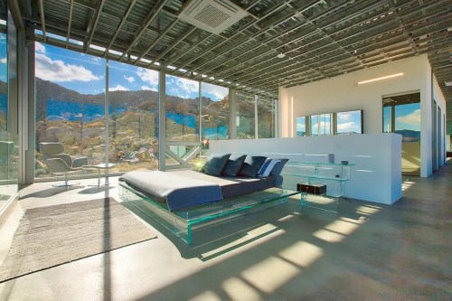 Modern bedroom with a glass bed frame, large windows, and a view of rocky mountains and blue skies.