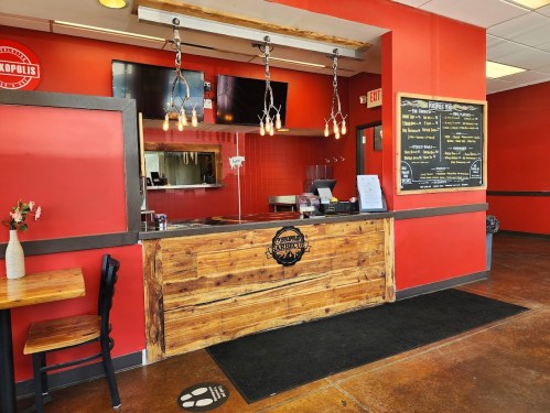 A cozy restaurant interior featuring a wooden counter, red walls, and a menu board. Tables are set for dining.