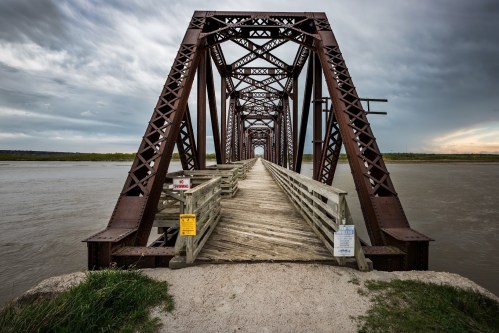 A rusty metal bridge spans a river, with wooden walkways and cloudy skies in the background.