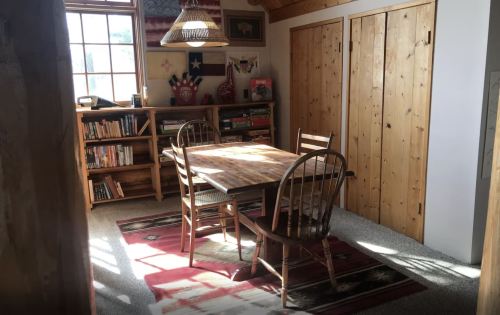 Cozy dining area with a wooden table, chairs, and a bookshelf, illuminated by sunlight through a window.