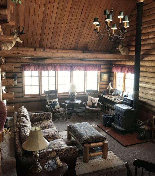 Cozy log cabin living room with a sofa, armchairs, a chandelier, and a wood stove, featuring rustic decor and large windows.