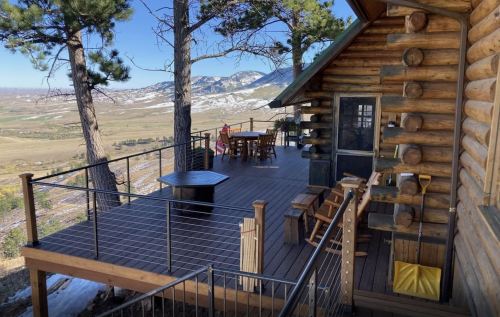 A wooden cabin deck with a table and chairs, surrounded by trees and mountains in the background.