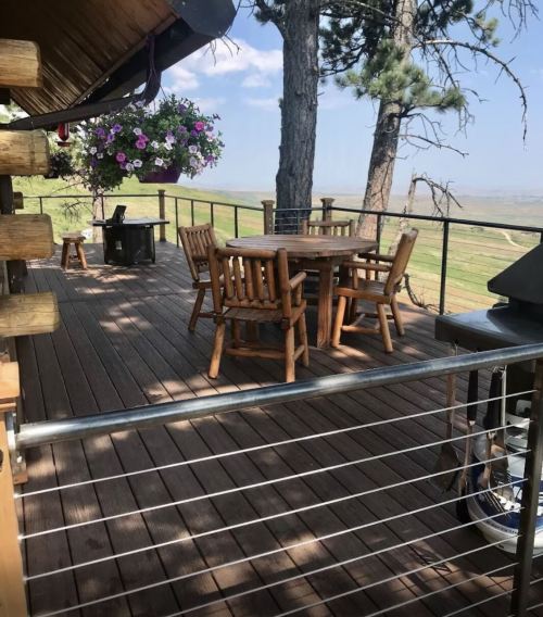 A wooden deck with a round table and chairs, surrounded by trees and a scenic view of rolling hills.