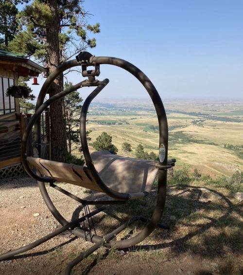 A hanging chair on a hillside, overlooking a vast valley and distant mountains under a clear blue sky.