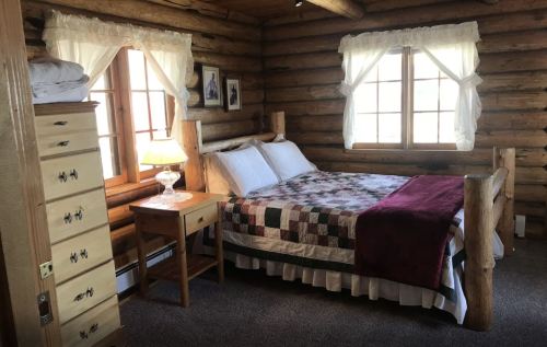 Cozy log cabin bedroom with a quilted bed, wooden furniture, and large windows letting in natural light.