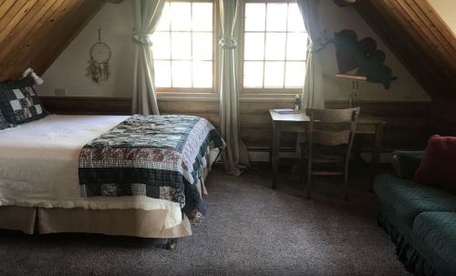 Cozy attic bedroom with a bed, desk, and seating area, featuring wooden beams and large windows.
