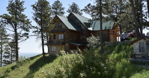 A log cabin with a green roof surrounded by tall trees and lush grass, overlooking a scenic view.